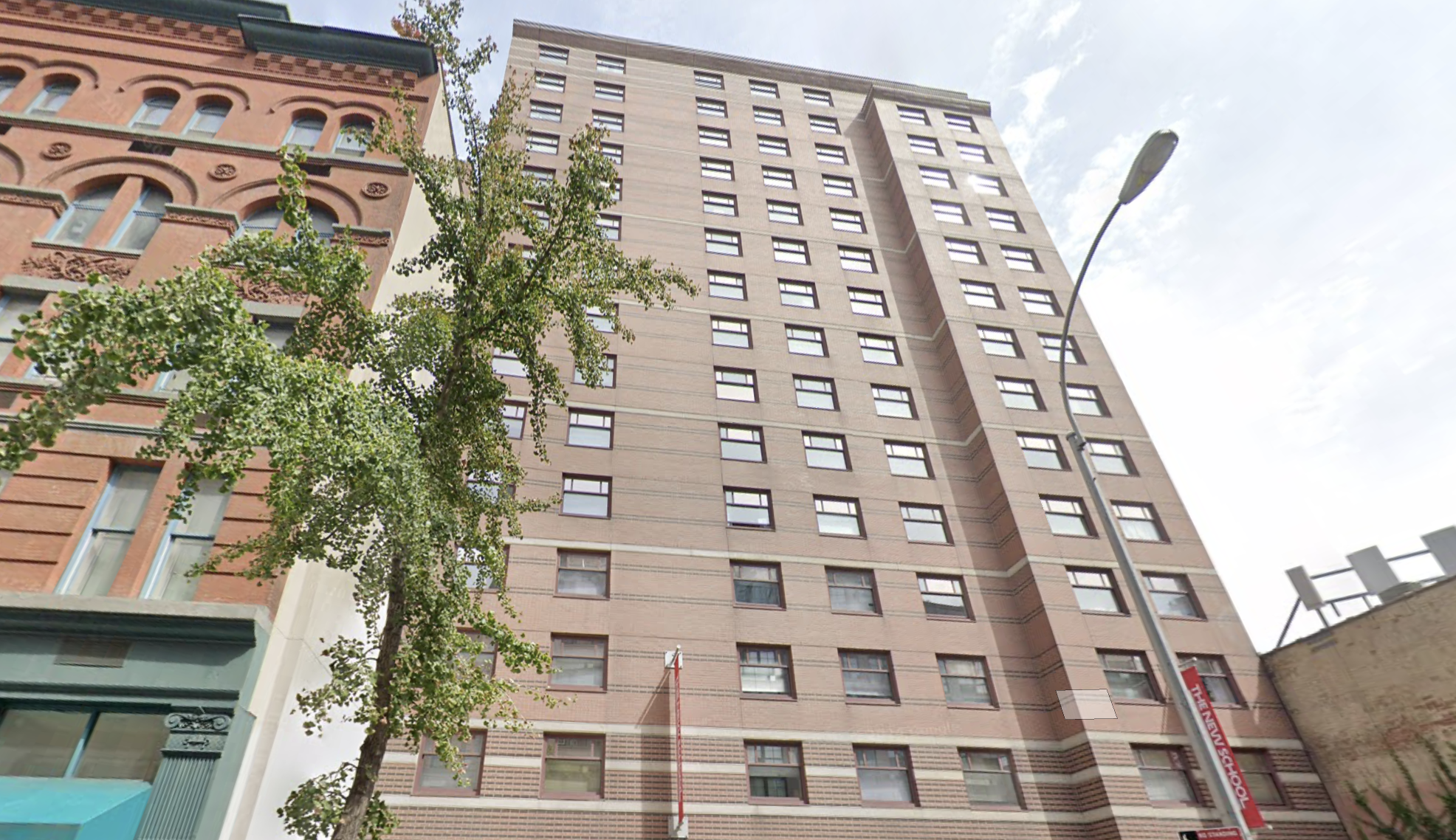 Tall residential building with uniform windows, red brick facade, adjacent to a historic structure, under a clear sky. A tree and streetlamp are visible.