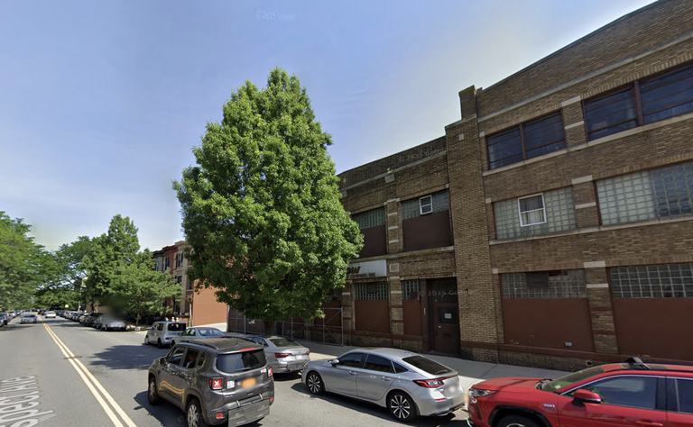 Street view with parked cars, a tall tree, and a brick industrial building under a clear blue sky.