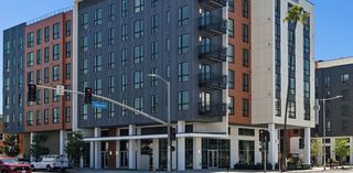 A mid-rise building at a city intersection, featuring large windows and balconies. Also visible are cars and traffic lights under a clear sky.