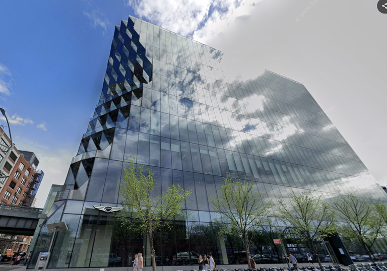 Modern glass building with geometric patterns, reflecting a partly cloudy sky. Trees and pedestrians are visible at the street level.