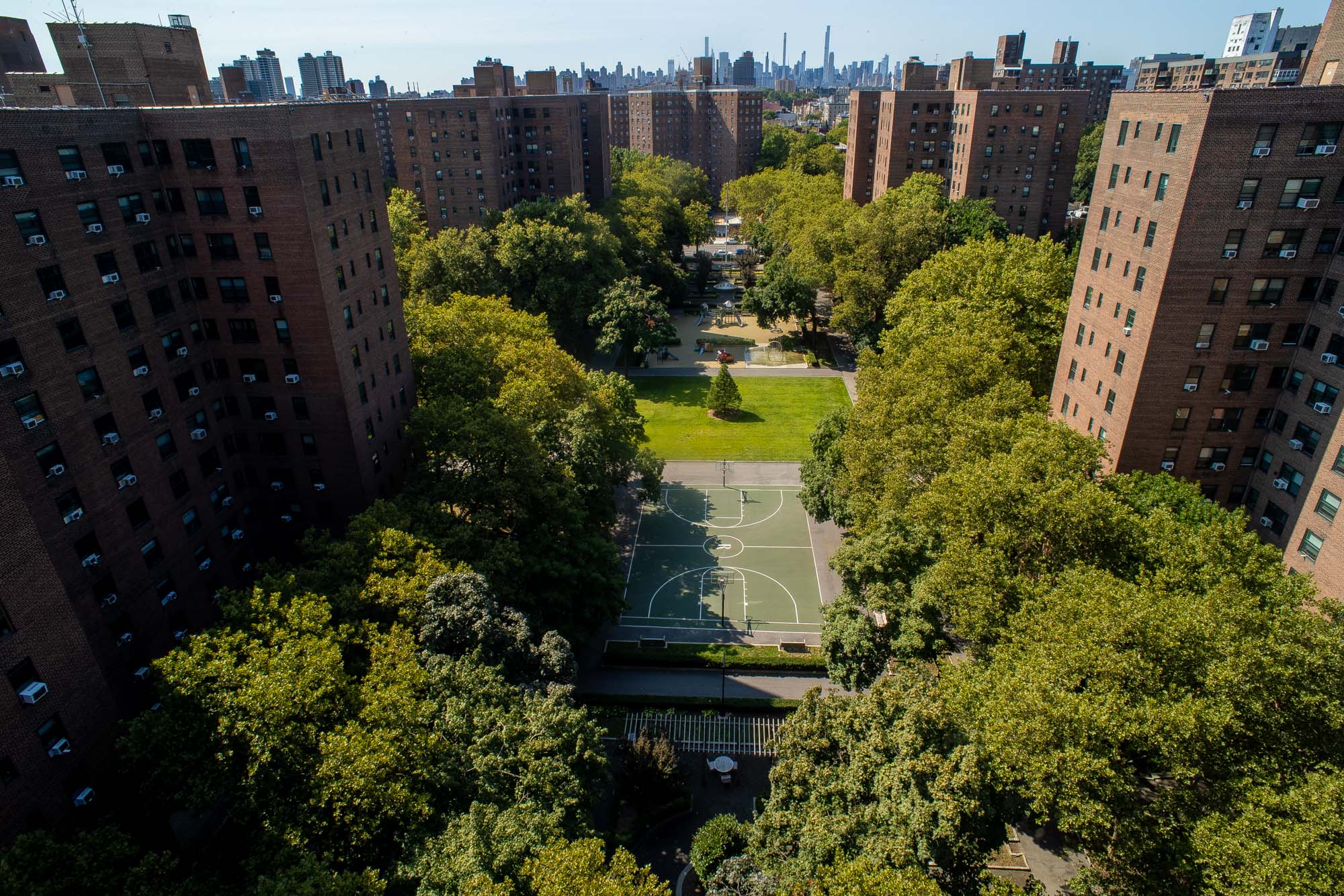 Elevated view of Riverton Square's courtyard surrounded by tall brick buildings, featuring green trees, a basketball court, playground, and distant city skyline.