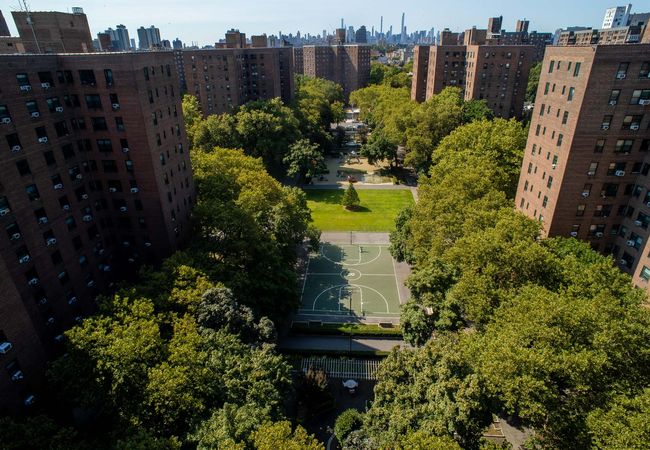 Elevated view of Riverton Square's courtyard surrounded by tall brick buildings, featuring green trees, a basketball court, playground, and distant city skyline.