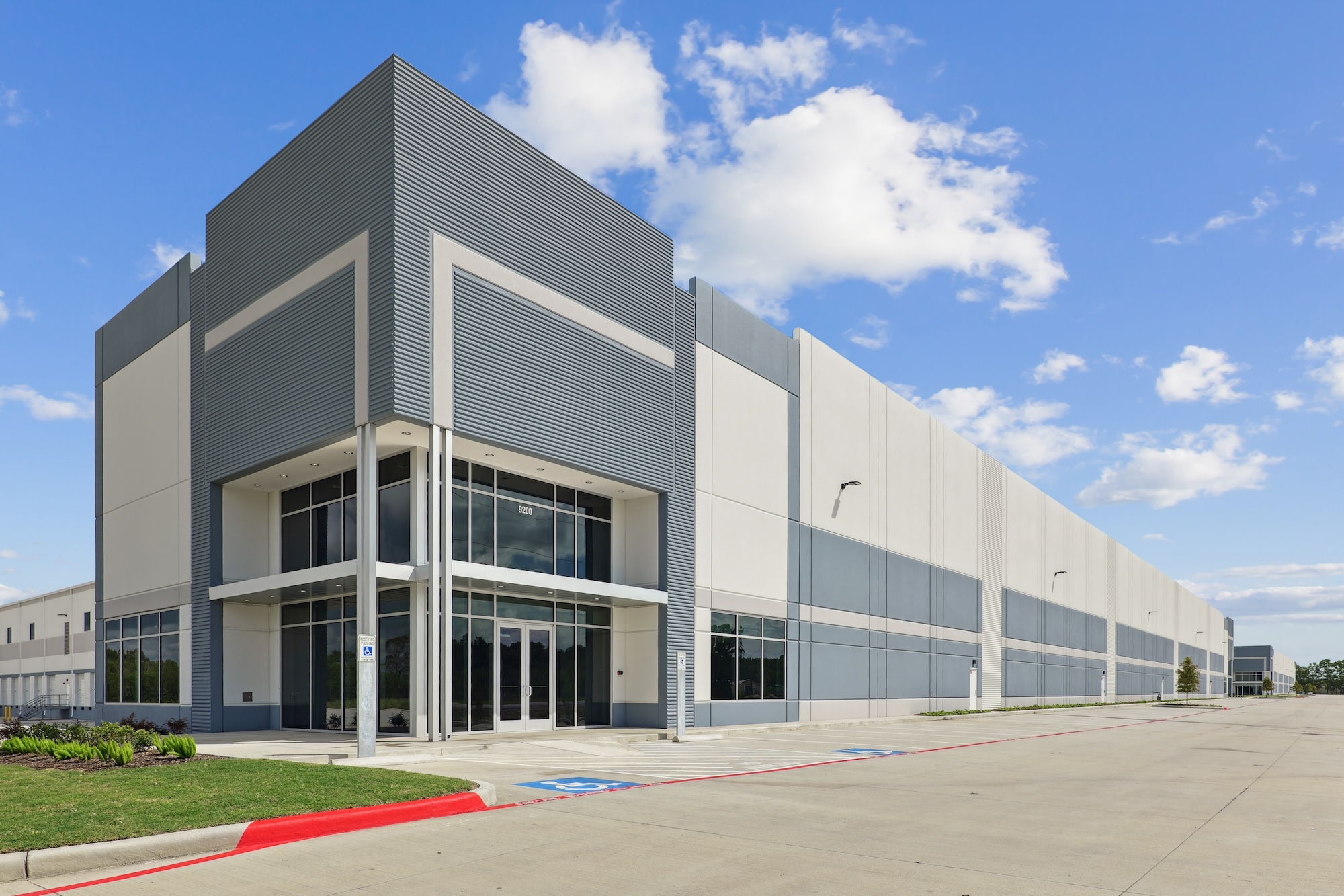 Modern industrial building with large glass entrance, gray and white facade, set against a blue sky with clouds.