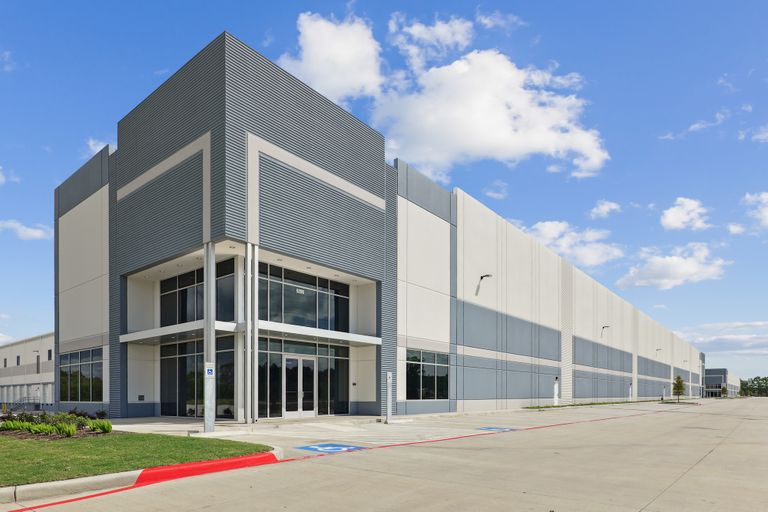 Modern industrial building with large glass entrance, gray and white facade, set against a blue sky with clouds.