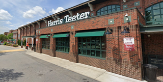 Brick facade of a grocery store with green awnings and a sign above. Sidewalk and parking lot visible under a partly cloudy sky.
