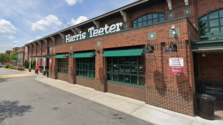 Brick facade of a grocery store with green awnings and a sign above. Sidewalk and parking lot visible under a partly cloudy sky.