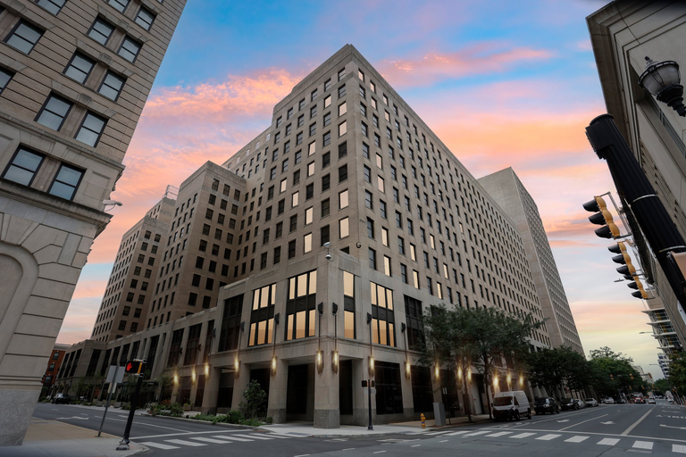A tall, L-shaped building under a colorful sunset sky, with empty streets and a traffic light in the foreground.