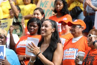 A diverse group of people in orange and yellow shirts gather outdoors, holding signs and smiling. A woman in front claps, holding papers.