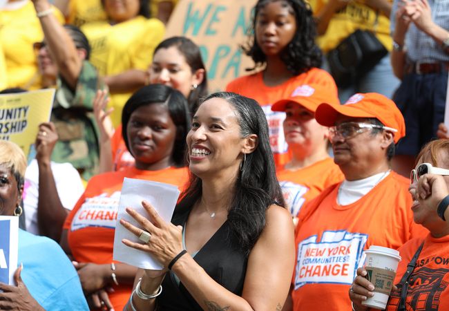 A diverse group of people in orange and yellow shirts gather outdoors, holding signs and smiling. A woman in front claps, holding papers.