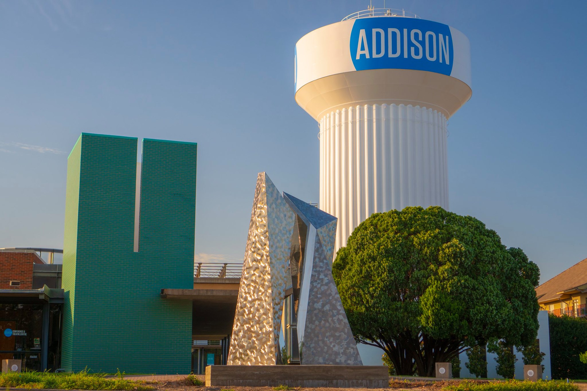 A water tower labeled "Addison" beside a modern sculpture and green building, with a large tree and clear blue sky.