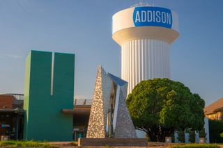 A water tower labeled "Addison" beside a modern sculpture and green building, with a large tree and clear blue sky.