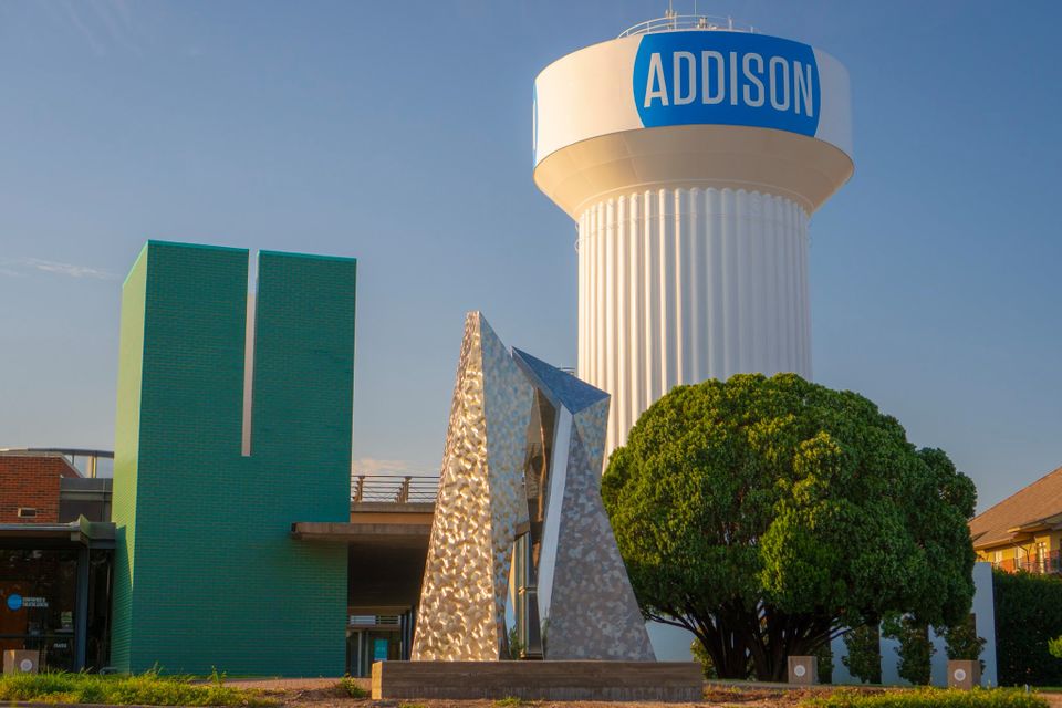 A water tower labeled "Addison" beside a modern sculpture and green building, with a large tree and clear blue sky.