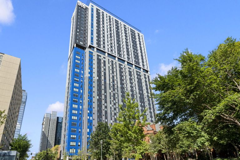 Tall modern high-rise building with a blue and gray facade, surrounded by trees and other buildings, under a clear blue sky.