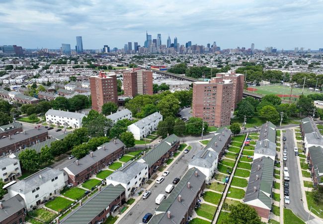 Aerial view of an urban neighborhood with rows of houses and tall apartment buildings, set against a city skyline under a cloudy sky.