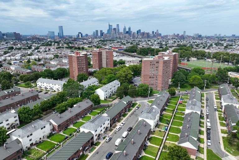 Aerial view of an urban neighborhood with rows of houses and tall apartment buildings, set against a city skyline under a cloudy sky.