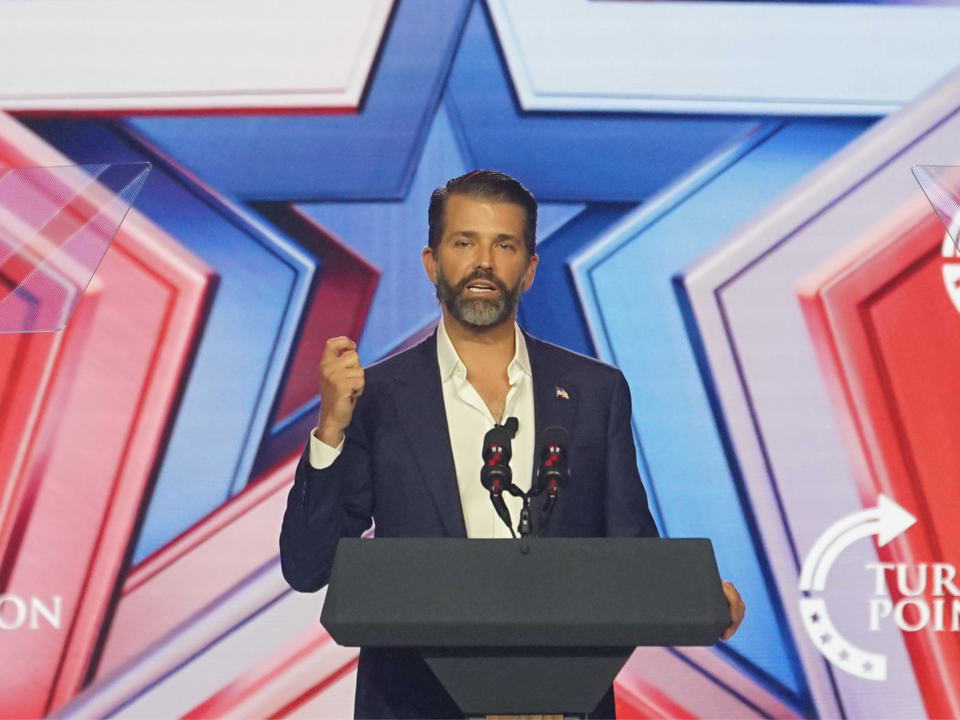 Donald Trump Jr. speaks at a podium with multiple microphones, against a backdrop featuring red and blue geometric patterns.