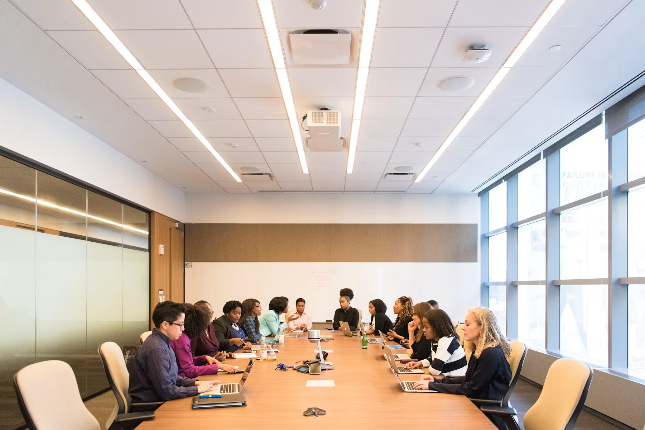 A group of 14 people sitting around a long conference table with laptops, engaged in discussion, in a modern office meeting room with large windows.