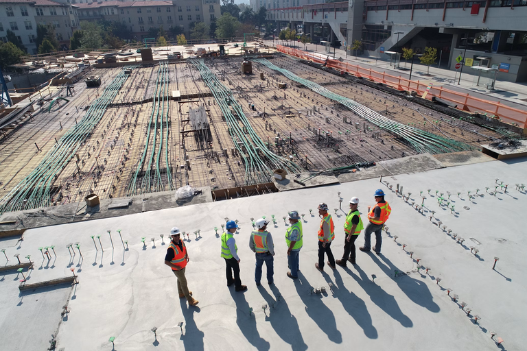 A group of construction workers wearing hard hats and safety vests stand on a concrete slab at a large building site, discussing the project.