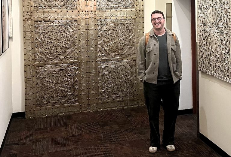 A person with glasses stands smiling in front of an intricately carved double door, wearing a brown jacket and carrying a backpack.