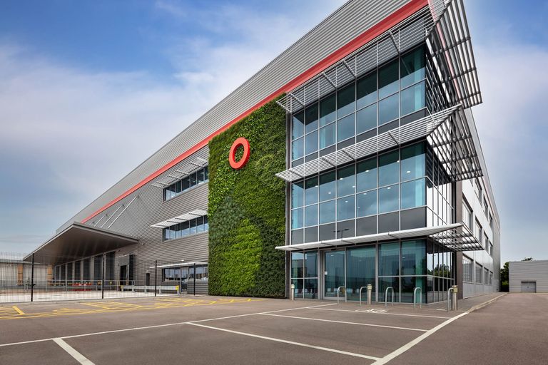 Modern industrial building with large windows, a living green wall, and a red circular logo on top, against a clear blue sky.