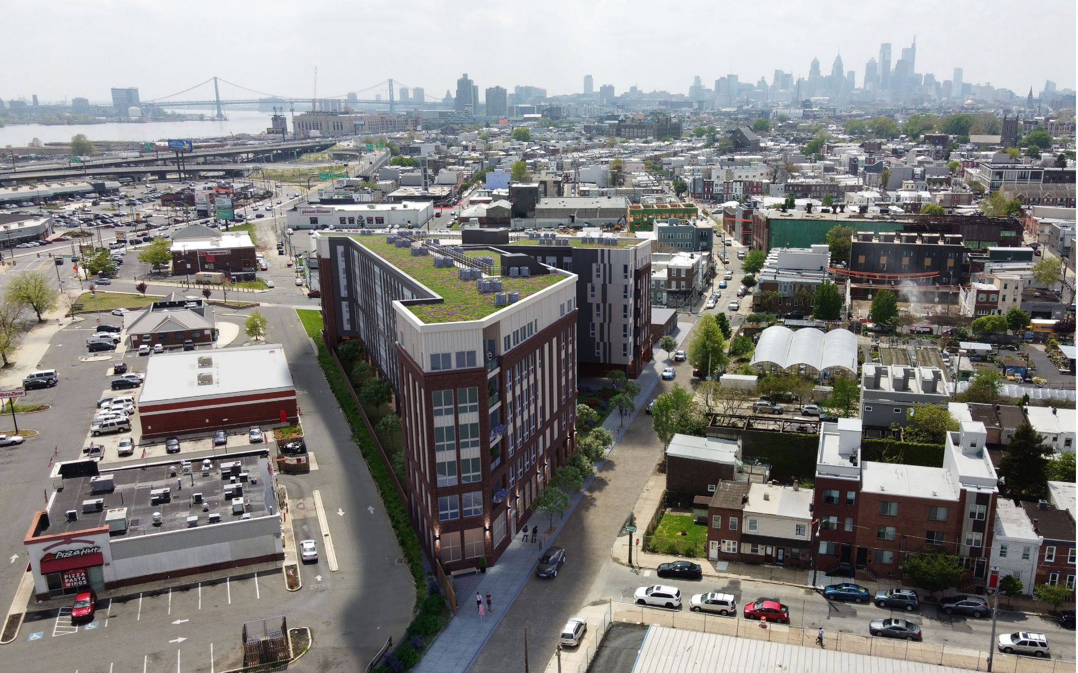 Aerial view of an urban area with buildings, a green-roofed structure, parked cars, and a distant city skyline near a river and bridge.