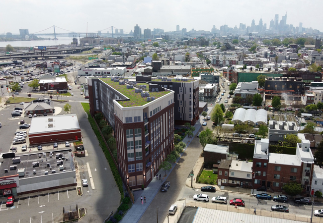 Aerial view of an urban area with buildings, a green-roofed structure, parked cars, and a distant city skyline near a river and bridge.