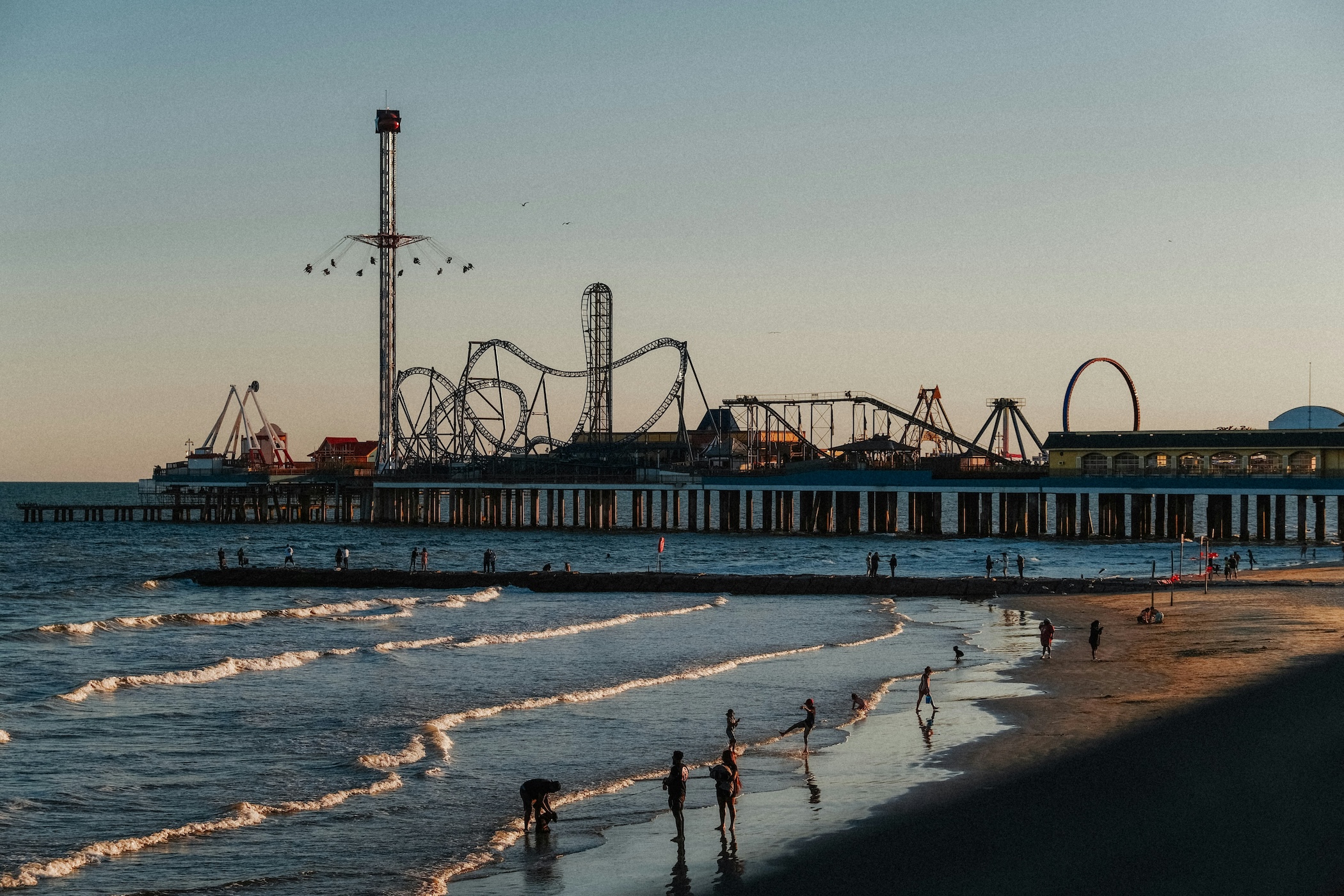 People on the beach near an amusement park with roller coasters and rides on a pier at sunset.