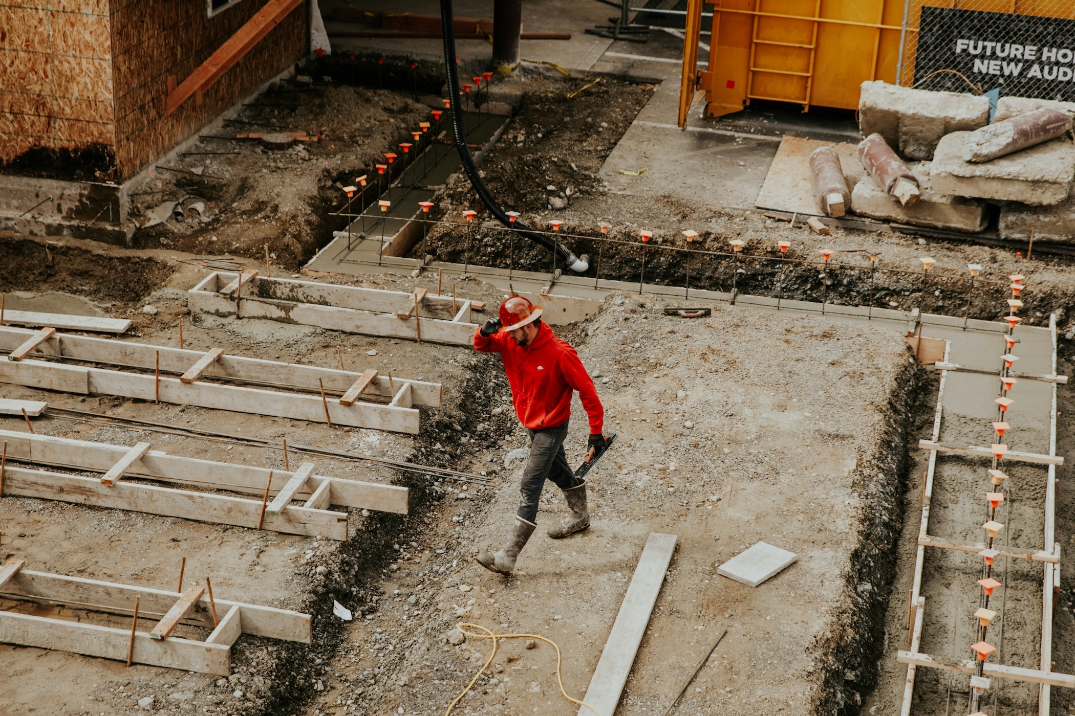 A construction worker in a red hoodie and hard hat walks at a construction site, surrounded by concrete forms and building materials.