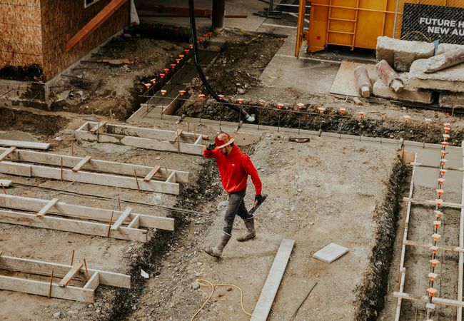 A construction worker in a red hoodie and hard hat walks at a construction site, surrounded by concrete forms and building materials.