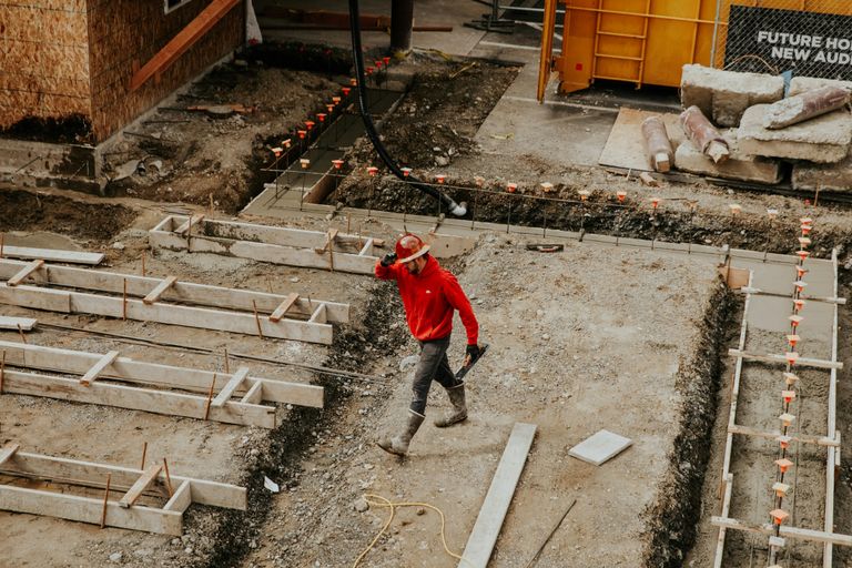 A construction worker in a red hoodie and hard hat walks at a construction site, surrounded by concrete forms and building materials.