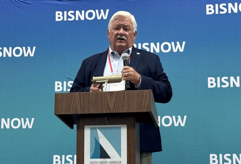Man speaking at a podium with a microphone against a blue "BISNOW" backdrop.