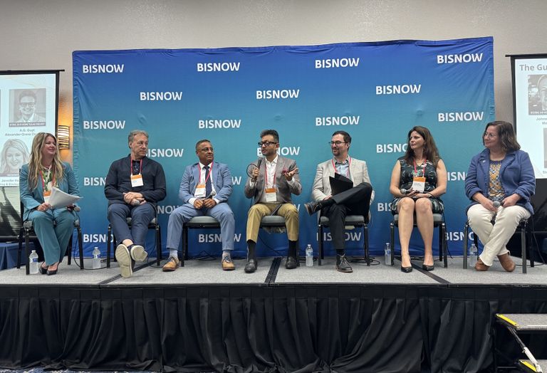 A panel of six people seated on a stage with BISNOW branding, engaging in a discussion, with two screens visible on either side.
