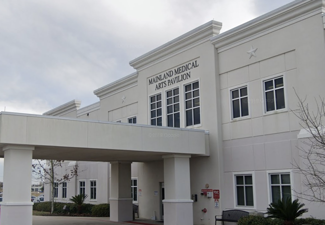 A beige, two-story building with a covered entrance labeled "Mainland Medical Arts Pavilion" against a cloudy sky, surrounded by trees and plants.