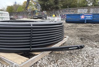 A large coil of black tubing sits on a wooden pallet at a construction site with gravel and dirt, surrounded by a graffitied wall and machinery.