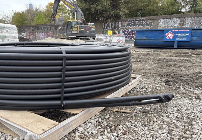 A large coil of black tubing sits on a wooden pallet at a construction site with gravel and dirt, surrounded by a graffitied wall and machinery.