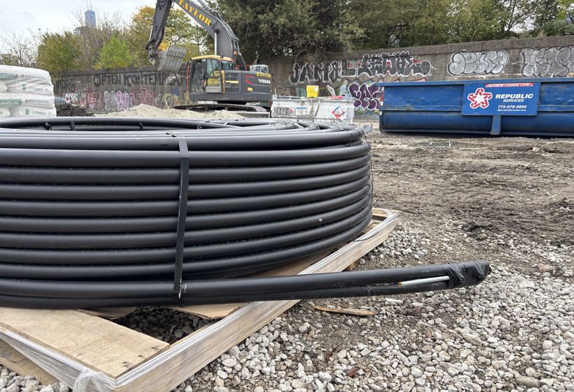 A large coil of black tubing sits on a wooden pallet at a construction site with gravel and dirt, surrounded by a graffitied wall and machinery.