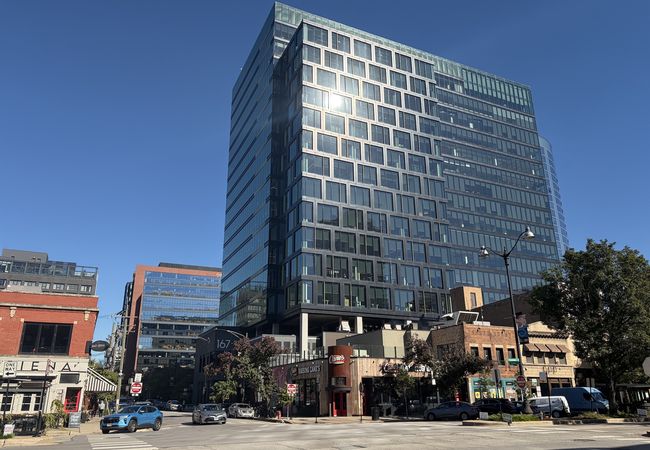 Urban street scene with modern glass high-rise, red brick buildings, and storefronts on a clear day. Cars and pedestrians visible in the foreground.