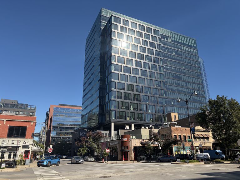 Urban street scene with modern glass high-rise, red brick buildings, and storefronts on a clear day. Cars and pedestrians visible in the foreground.