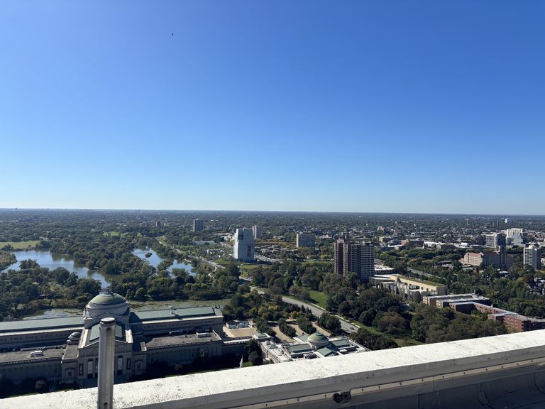 A panoramic view of a city with green spaces, lakes, and buildings, under a clear blue sky.