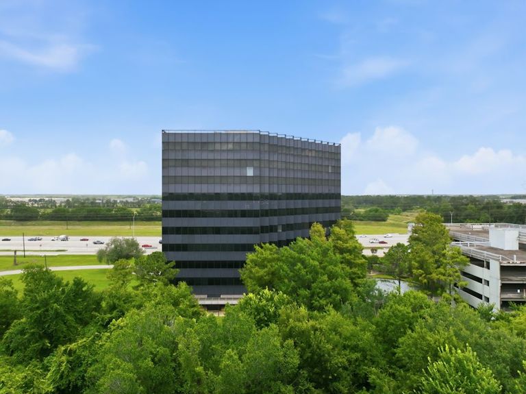 A modern, glass-paneled office building surrounded by lush green trees, with a highway and parked cars in the background under a clear blue sky.