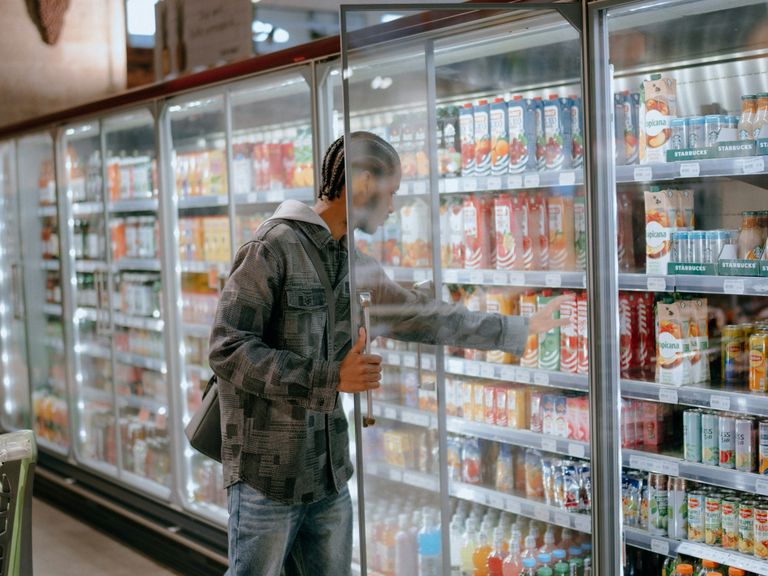 Person in a grocery store reaching into a refrigerated display filled with various bottled beverages, holding the door open.