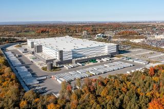 Aerial view of a large industrial warehouse surrounded by parking lots and autumn trees under a clear blue sky.