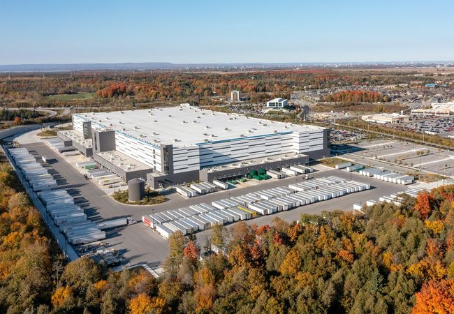 Aerial view of a large industrial warehouse surrounded by parking lots and autumn trees under a clear blue sky.