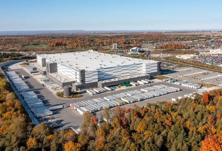Aerial view of a large industrial warehouse surrounded by parking lots and autumn trees under a clear blue sky.