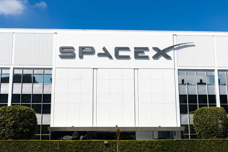 SpaceX building facade with large logo, blue sky, and neatly trimmed bushes in front.