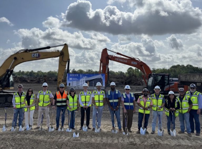 A group of people in safety vests and helmets holding shovels at a construction site with excavators in the background.
