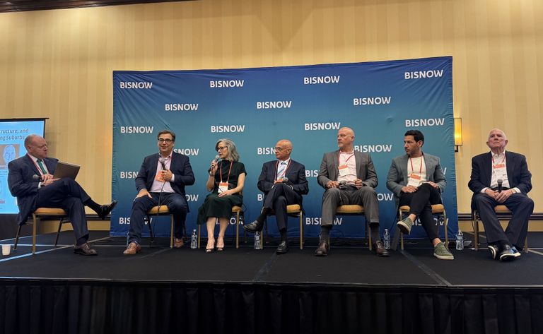 Seven panelists sit on a stage beneath a Bisnow banner, engaging in a discussion. One panelist speaks into a microphone.