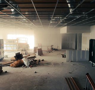 Large unfinished room with exposed ceiling grid, scattered construction materials, and a partially installed wall. Dust and light fill the space.