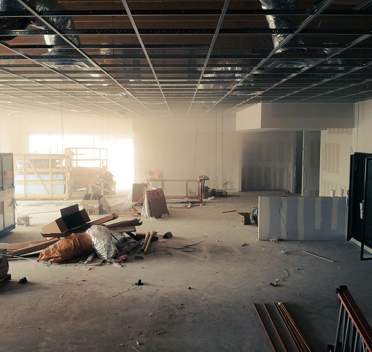 Large unfinished room with exposed ceiling grid, scattered construction materials, and a partially installed wall. Dust and light fill the space.