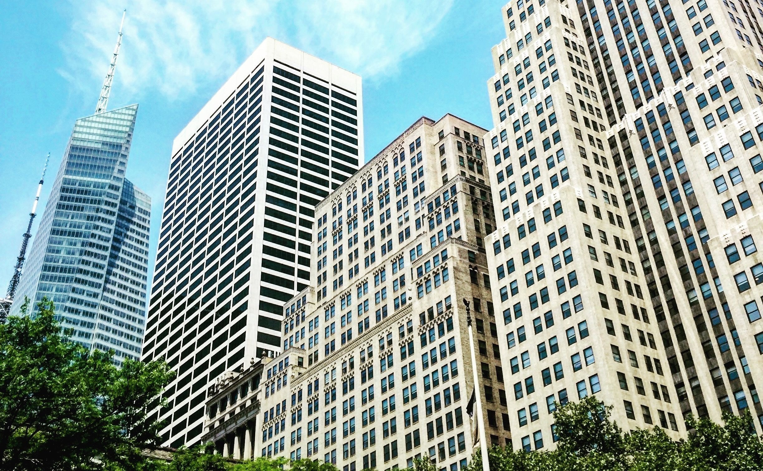 Tall city skyscrapers under a bright blue sky with scattered clouds, surrounded by green trees.